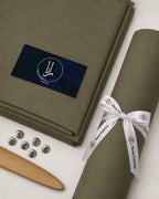 Olive green men's suiting fabric roll with Rana Fabrics ribbon, buttons, and wooden tool for formal wear